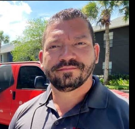 Man with dark hair and beard wearing a dark polo shirt, photographed outdoors with a red vehicle and palm trees in the background