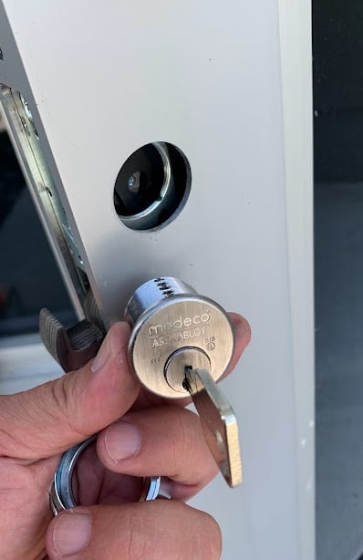 Hand holding a brass deadbolt lock with key inserted into a white door with peephole