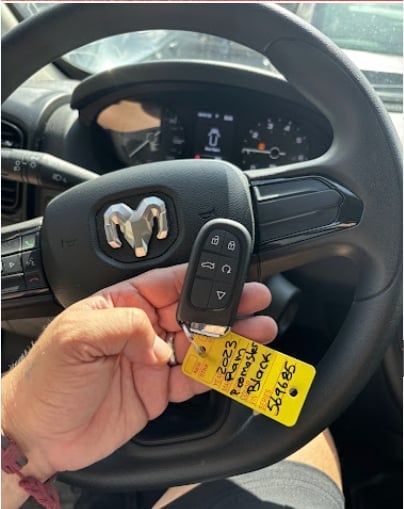 Hand holding a car key fob with a yellow price tag in front of a steering wheel inside a vehicle