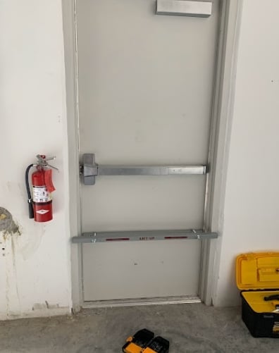 Emergency exit door with push bar, red fire extinguisher on left wall, and yellow toolbox on right in concrete room